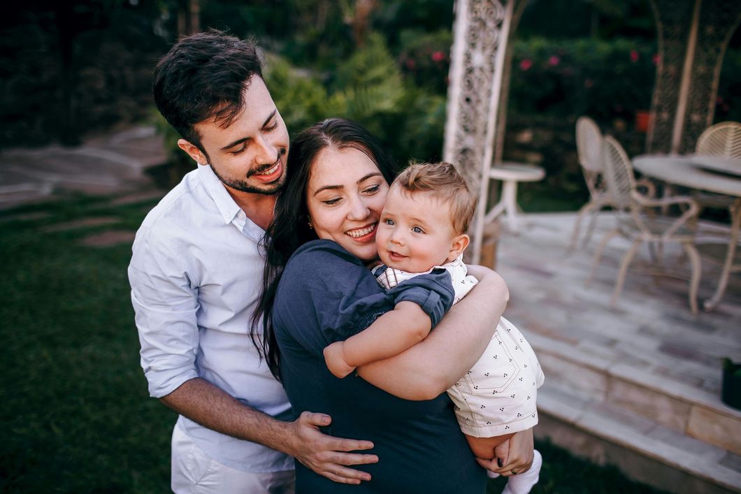 Two adults and a child hugging outdoors on a wooden deck, with chairs and greenery in the background