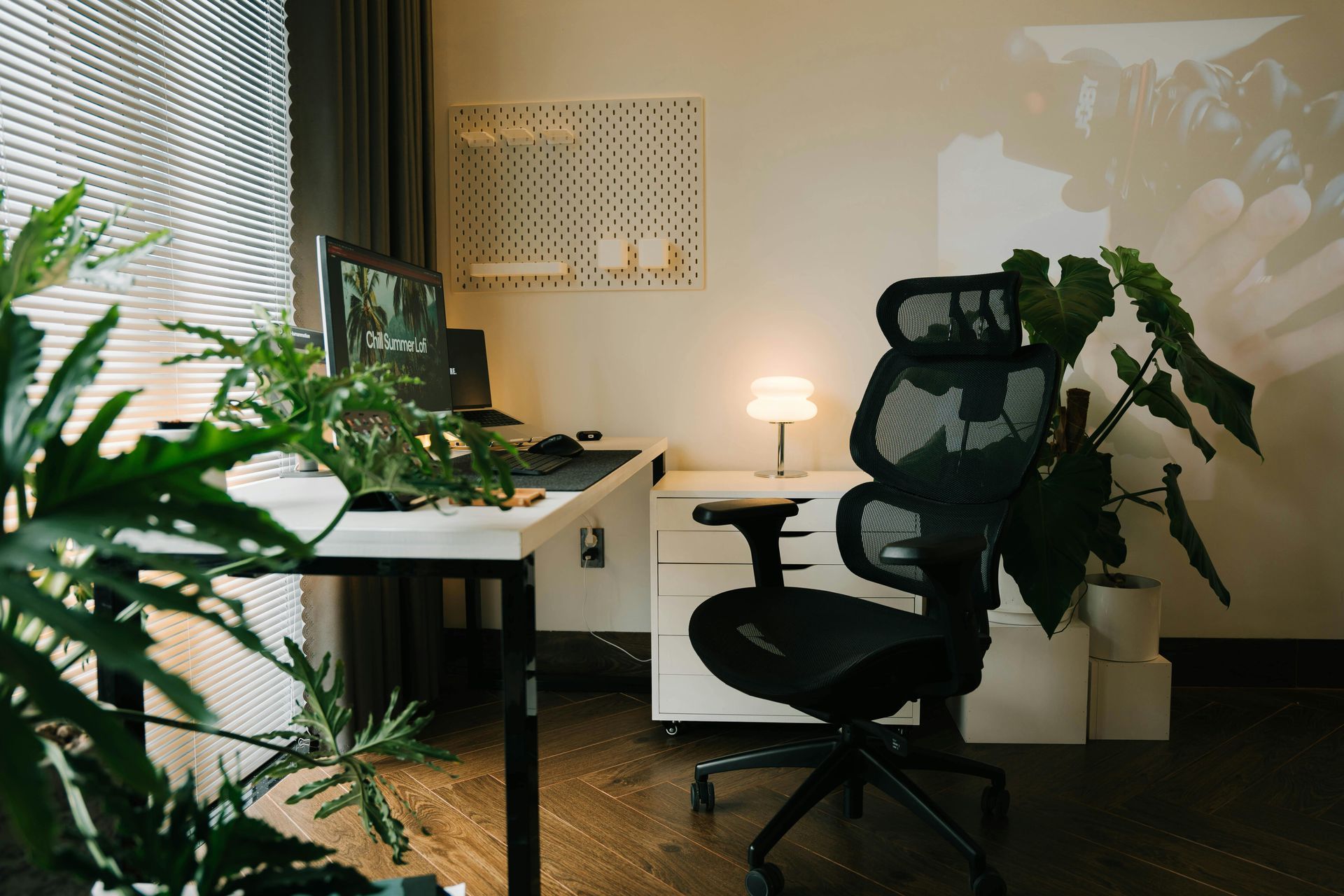 Modern home office with two desks, black ergonomic chair, plants, and a lit desk lamp by a window