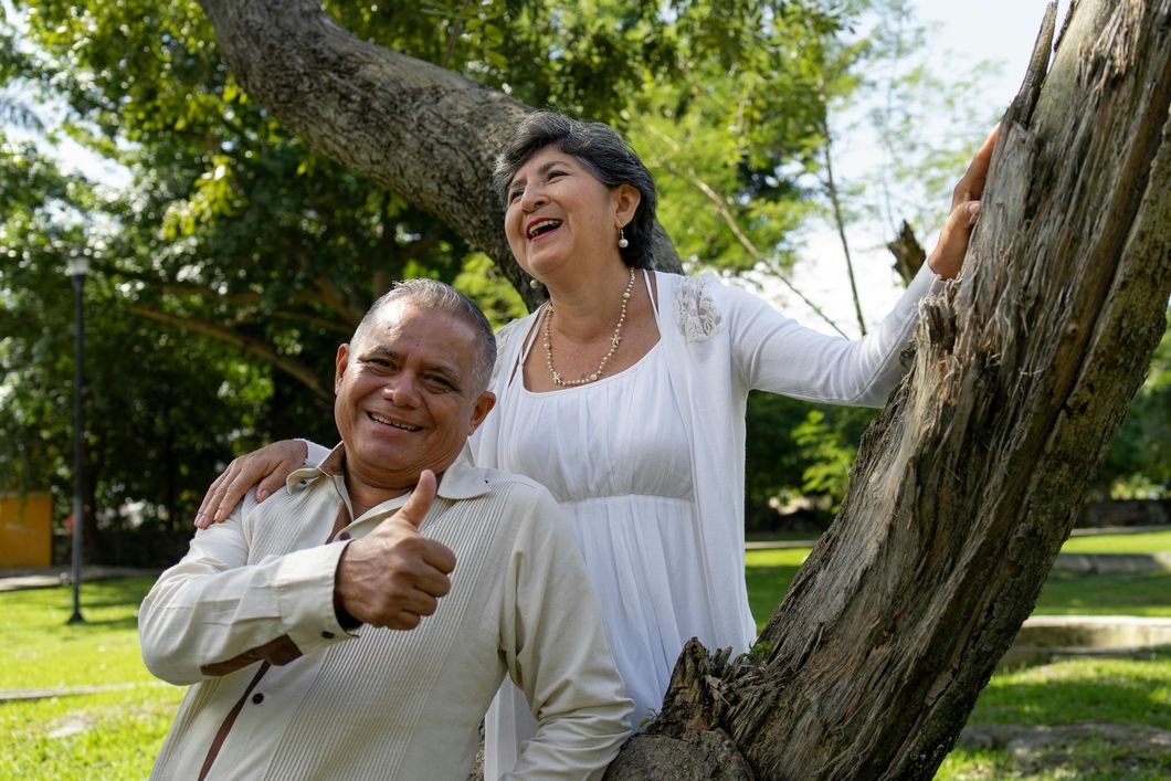 Two people smiling outdoors by a tree, one giving a thumbs-up, in a sunny park