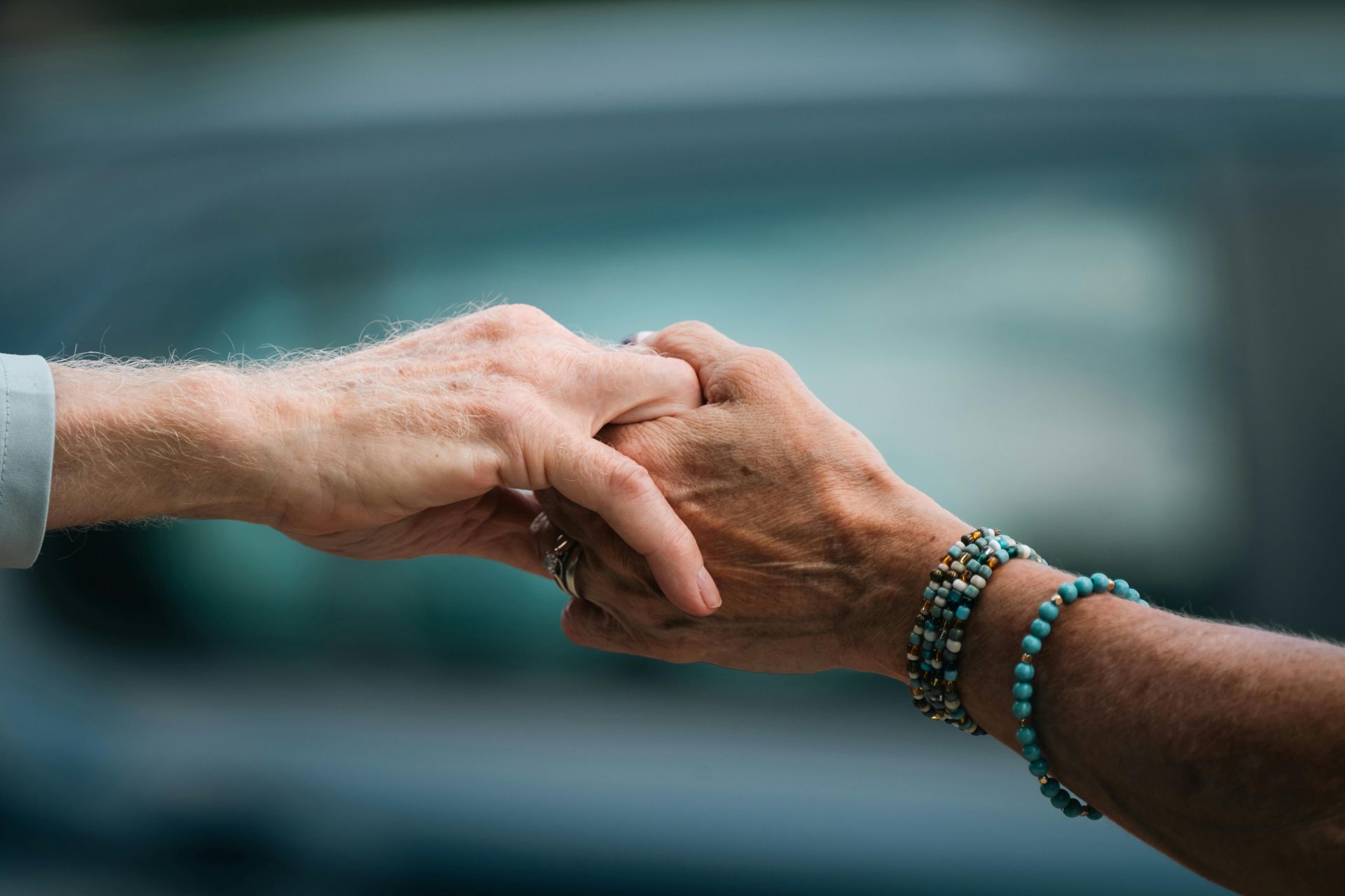 Two hands clasped in a handshake, one wearing beaded bracelets, against a blurred car background