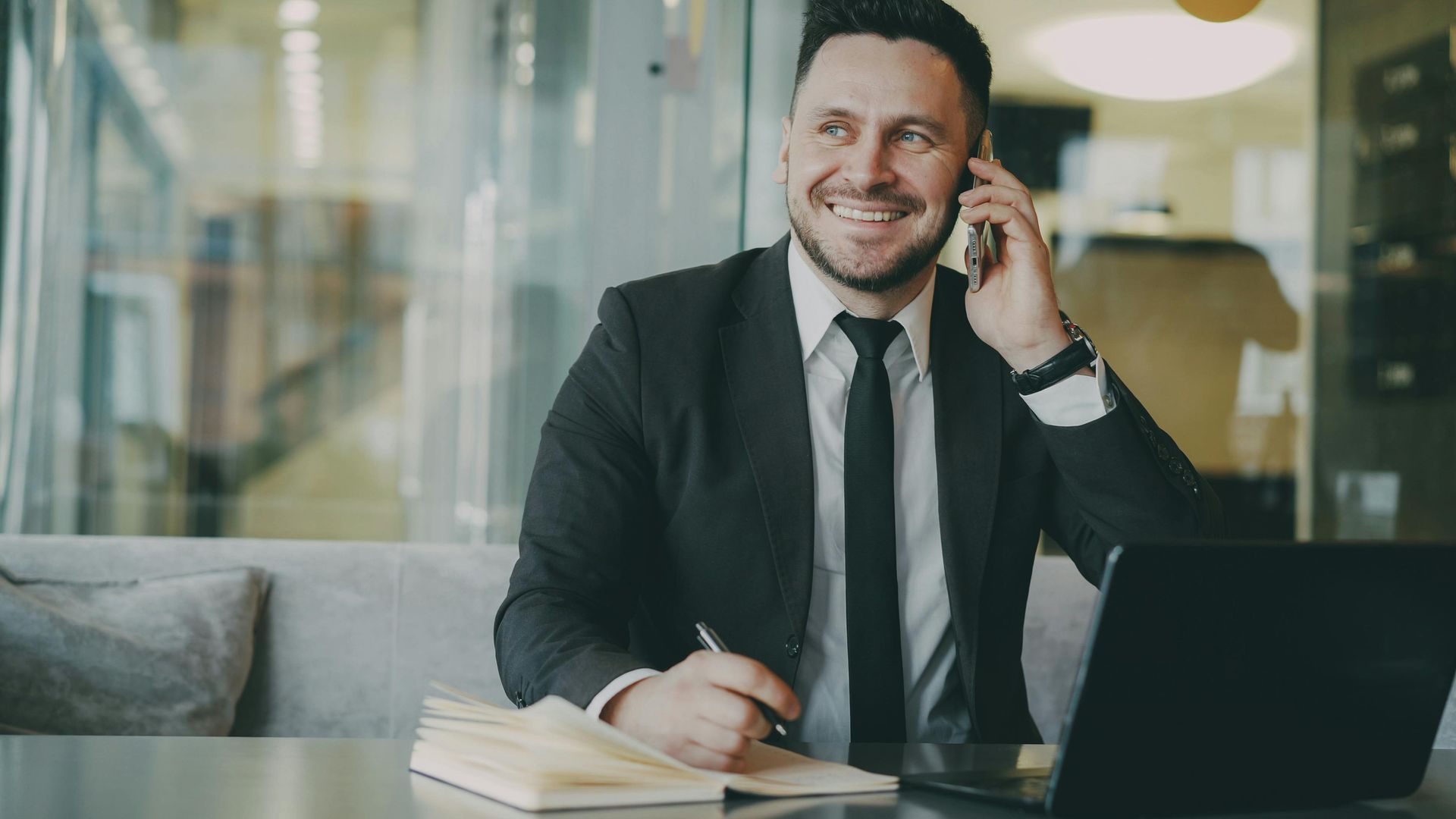 Man in a suit smiling while talking on a phone at a desk with a laptop and notebook in an office
