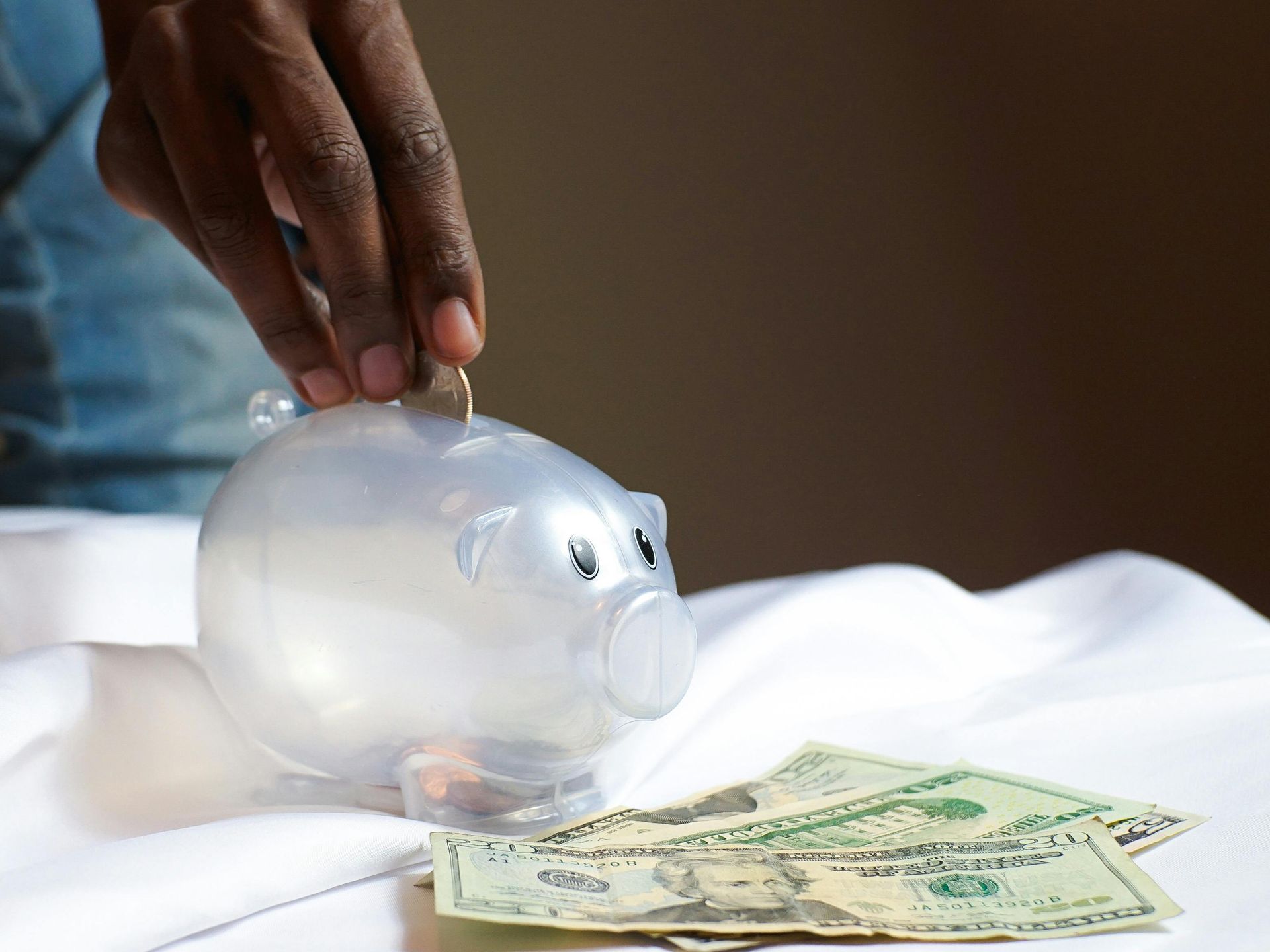 Hand placing a coin into a clear piggy bank beside scattered dollar bills
