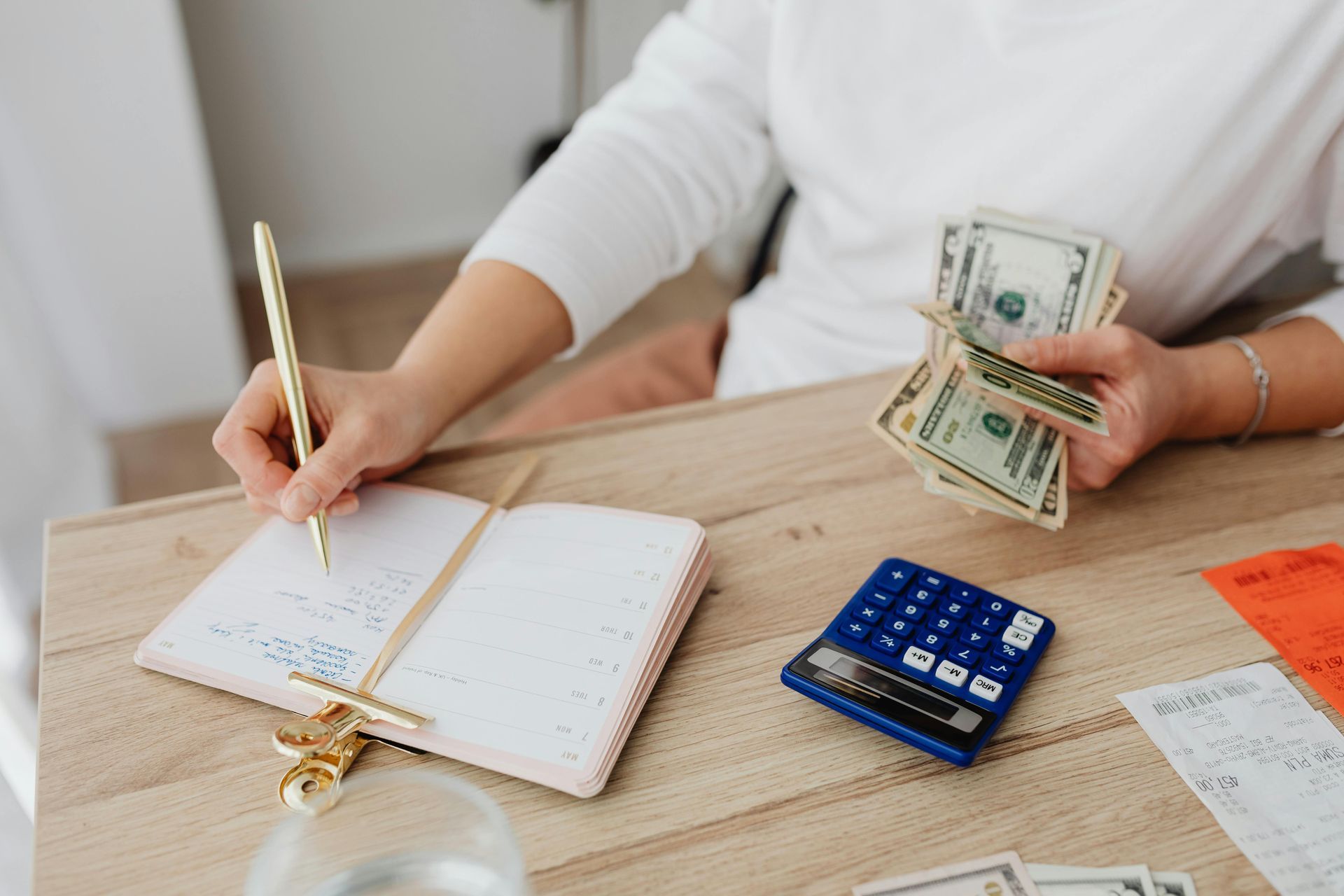 Person writing in a notebook beside cash and a blue calculator on a wooden desk.
