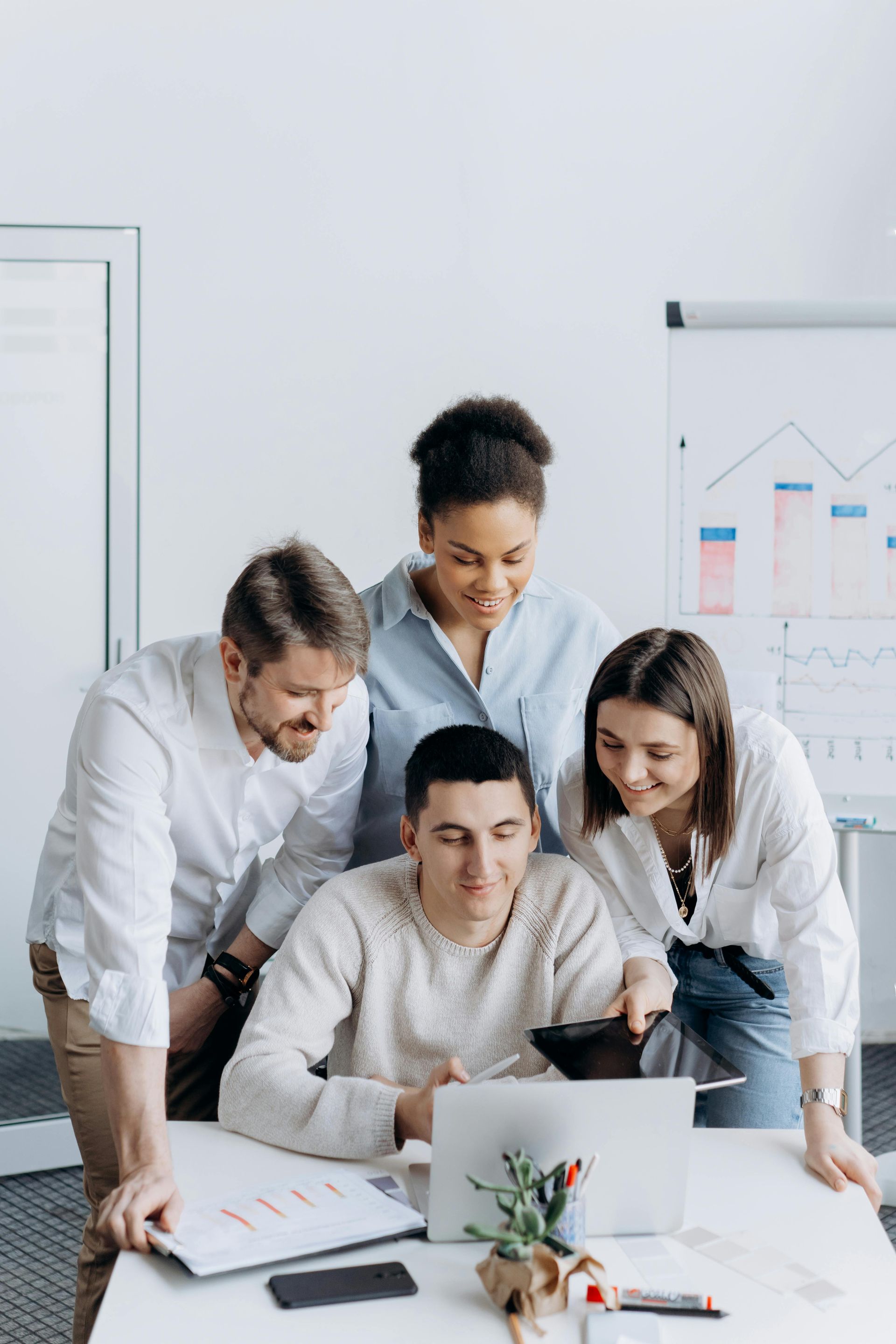 Four coworkers gathered around a laptop in a bright office, reviewing charts and documents together.