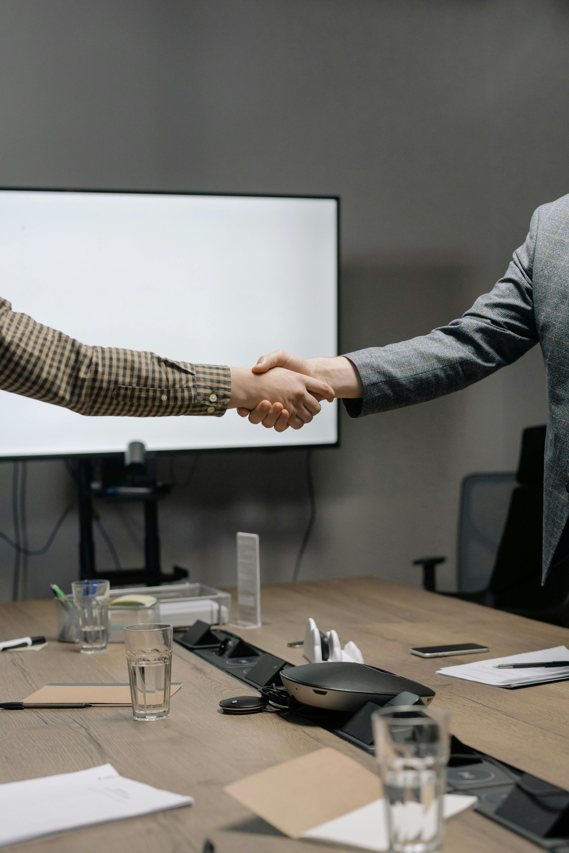 Two people shaking hands across a conference table with a presentation screen in the background