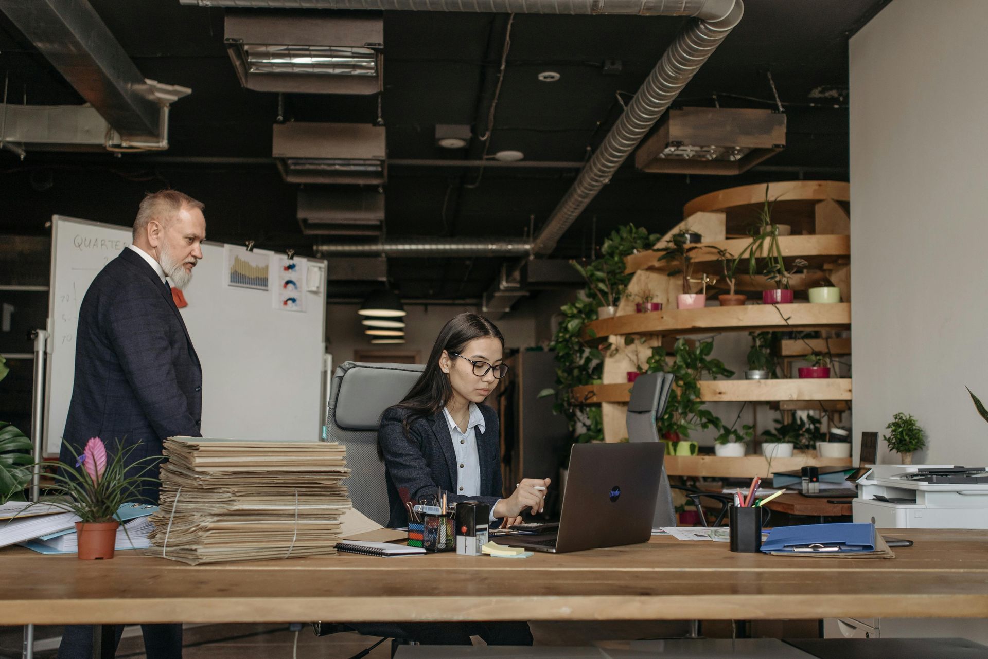 Two people in an office, one working at a laptop while another stands nearby, with shelves and plants in the background
