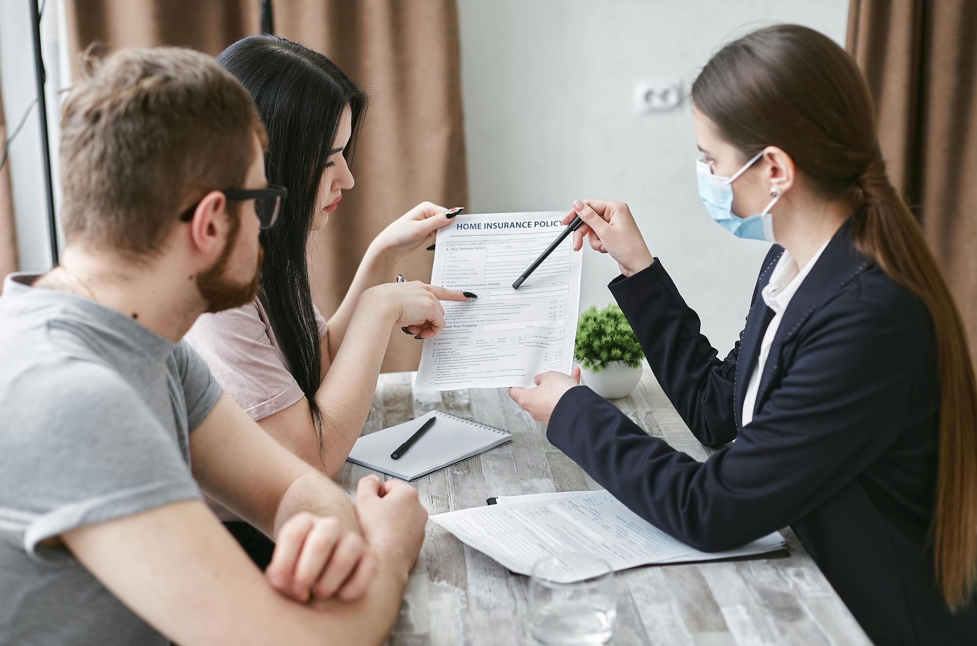 People in a meeting reviewing documents at a table, one wearing a face mask and pointing at a paper.