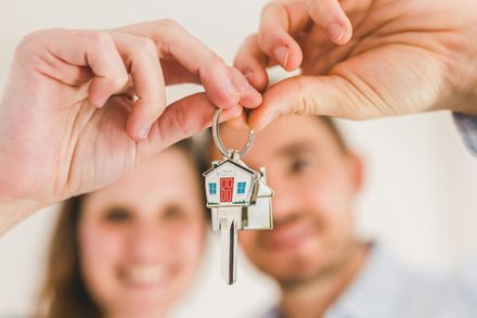 Hands holding a house key with a house-shaped keychain, with two smiling people blurred in the background