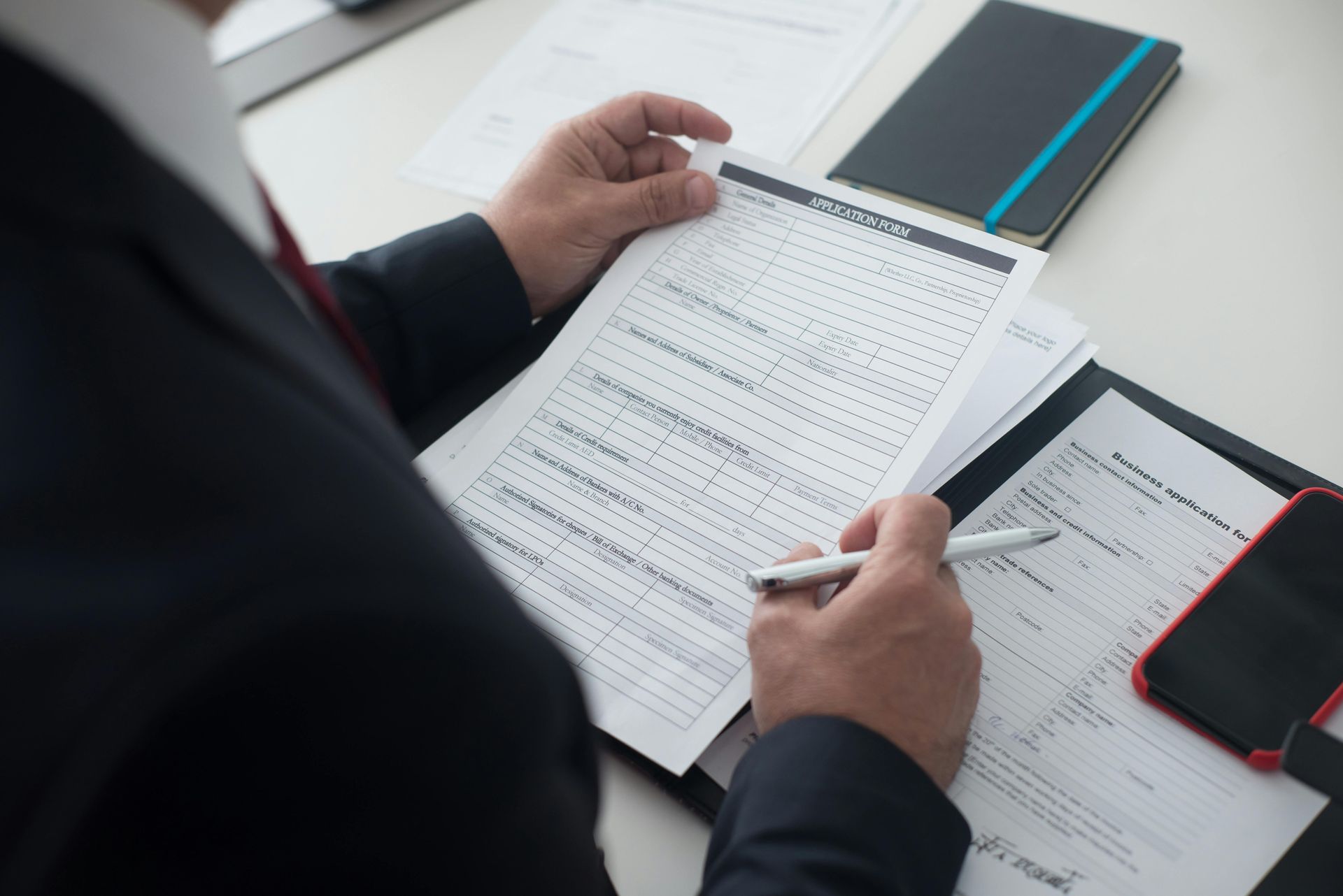 Person reviewing printed documents at a desk with a pen, notebook, and red phone nearby
