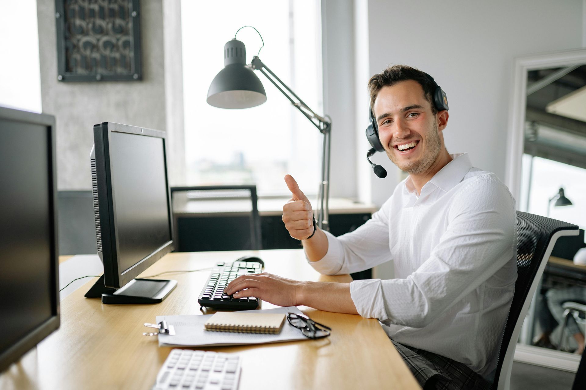 Smiling office worker wearing a headset at a desk, giving a thumbs-up beside a computer and lamp.