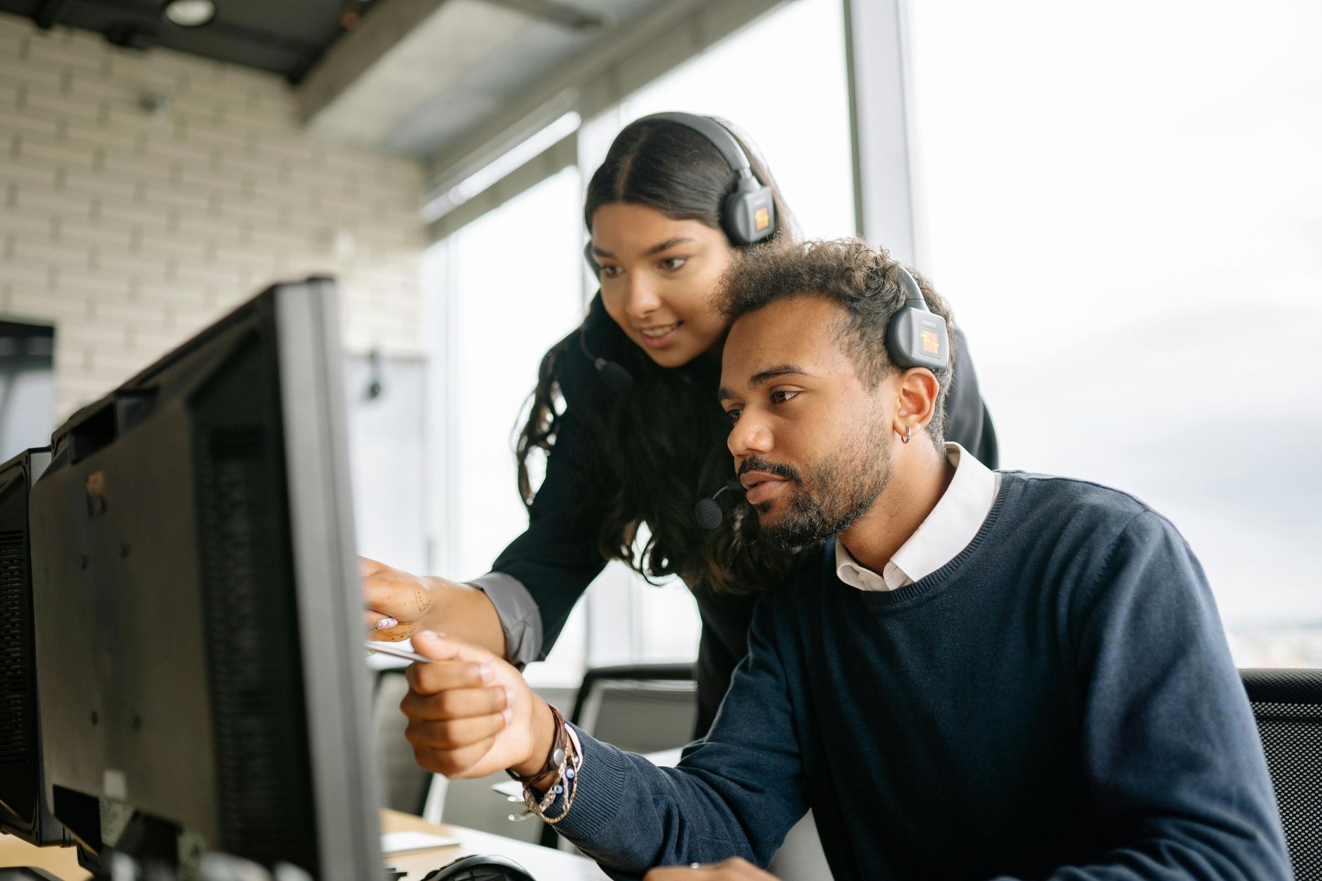 Two coworkers collaborate at a desktop computer in an office, one pointing at the screen.