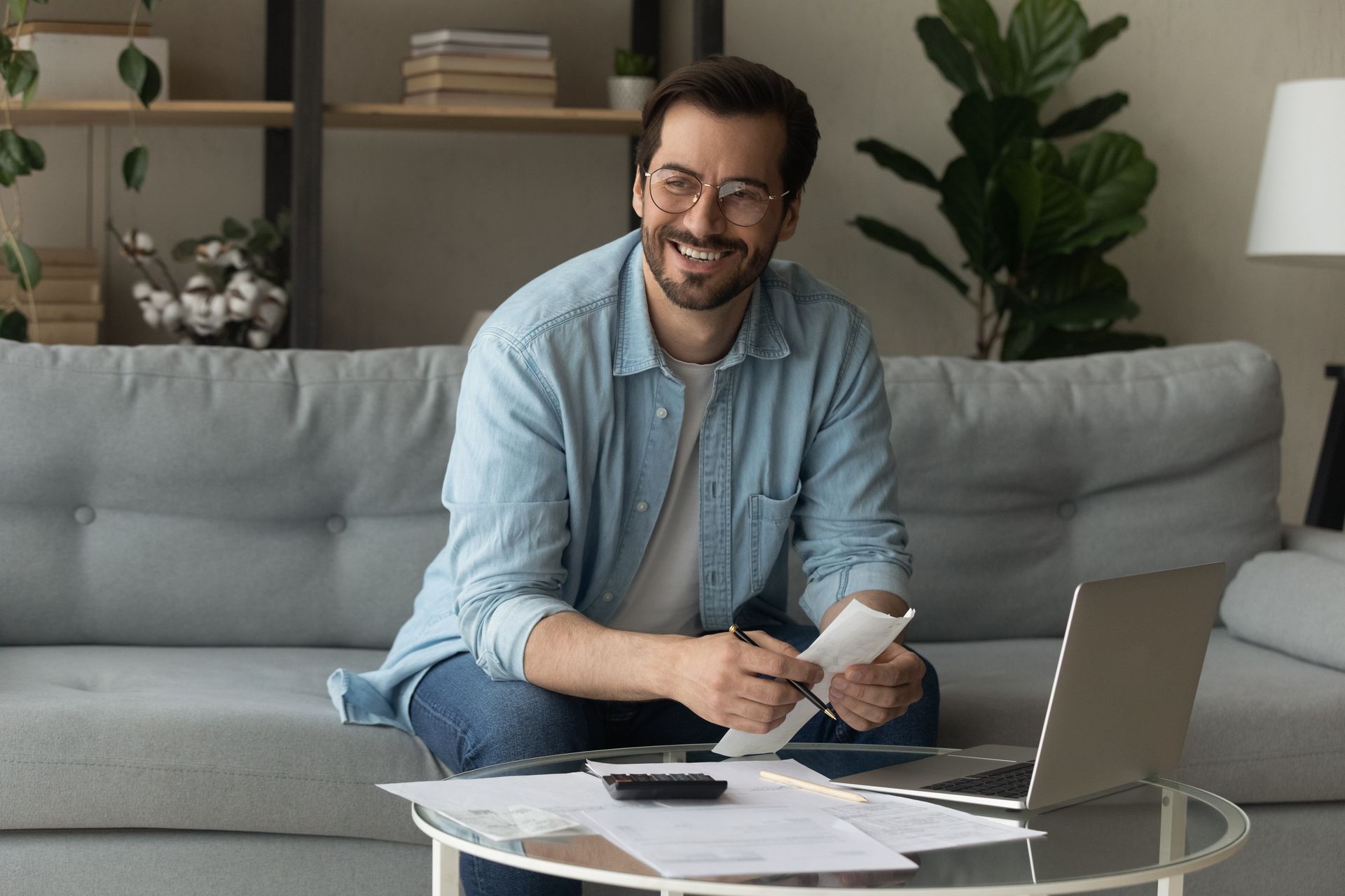 Man smiling on a couch, holding a card beside papers and a laptop on a glass coffee table.