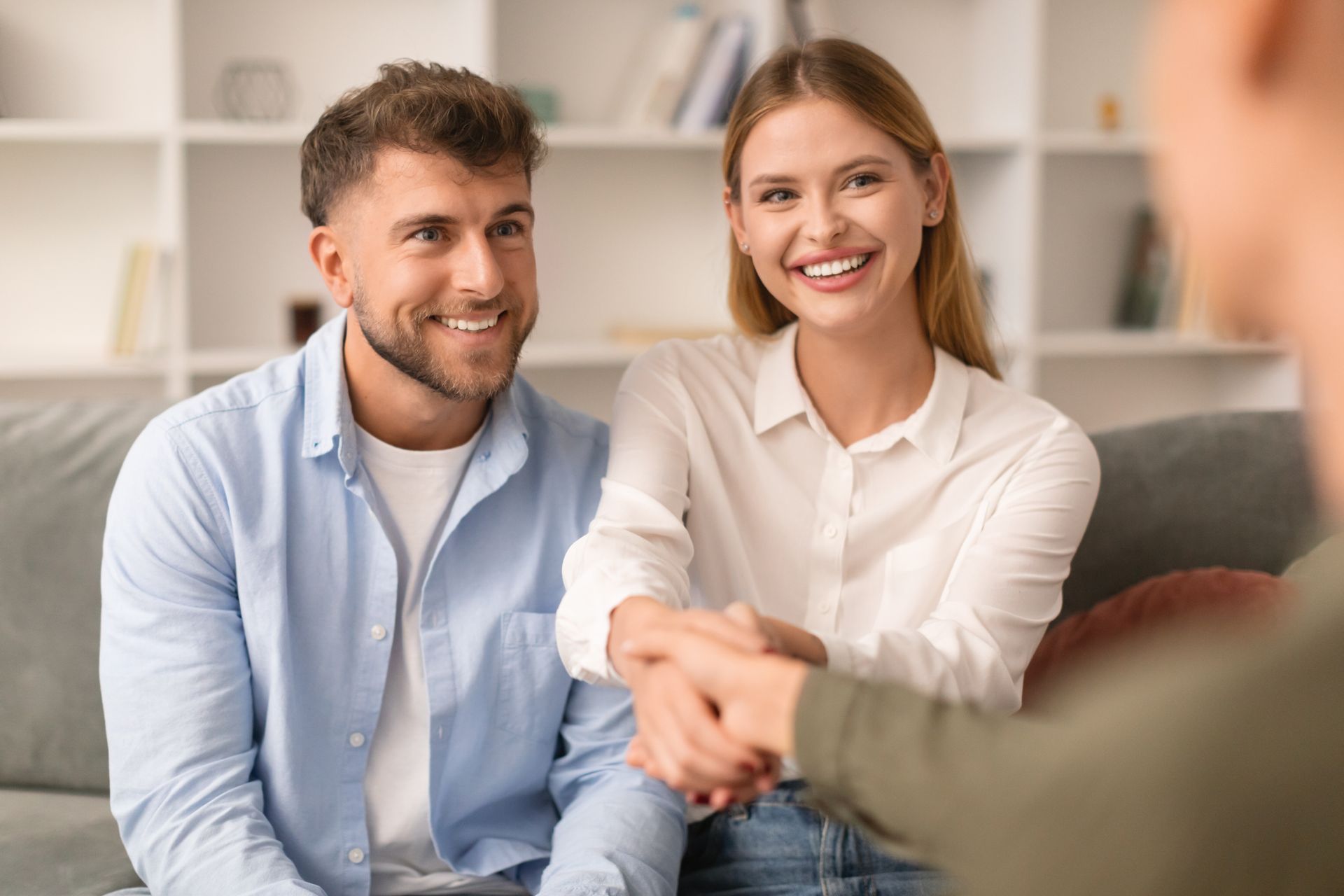 Two smiling people shaking hands in a bright living room, with a blurred third person in the foreground