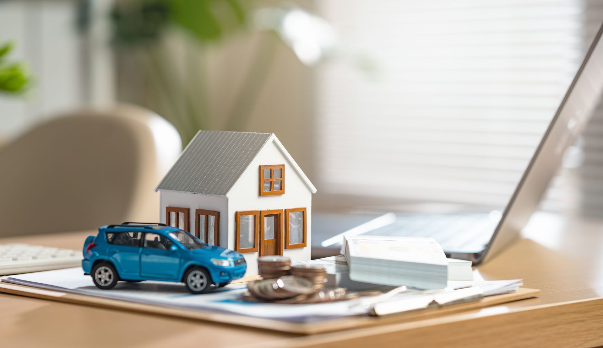 Toy house, blue car, and paperwork on a desk beside a laptop, suggesting home insurance or real estate.