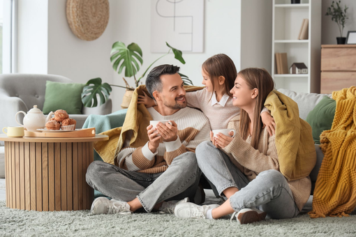 Three people sit on a living room rug, smiling and sharing popcorn in a cozy home.