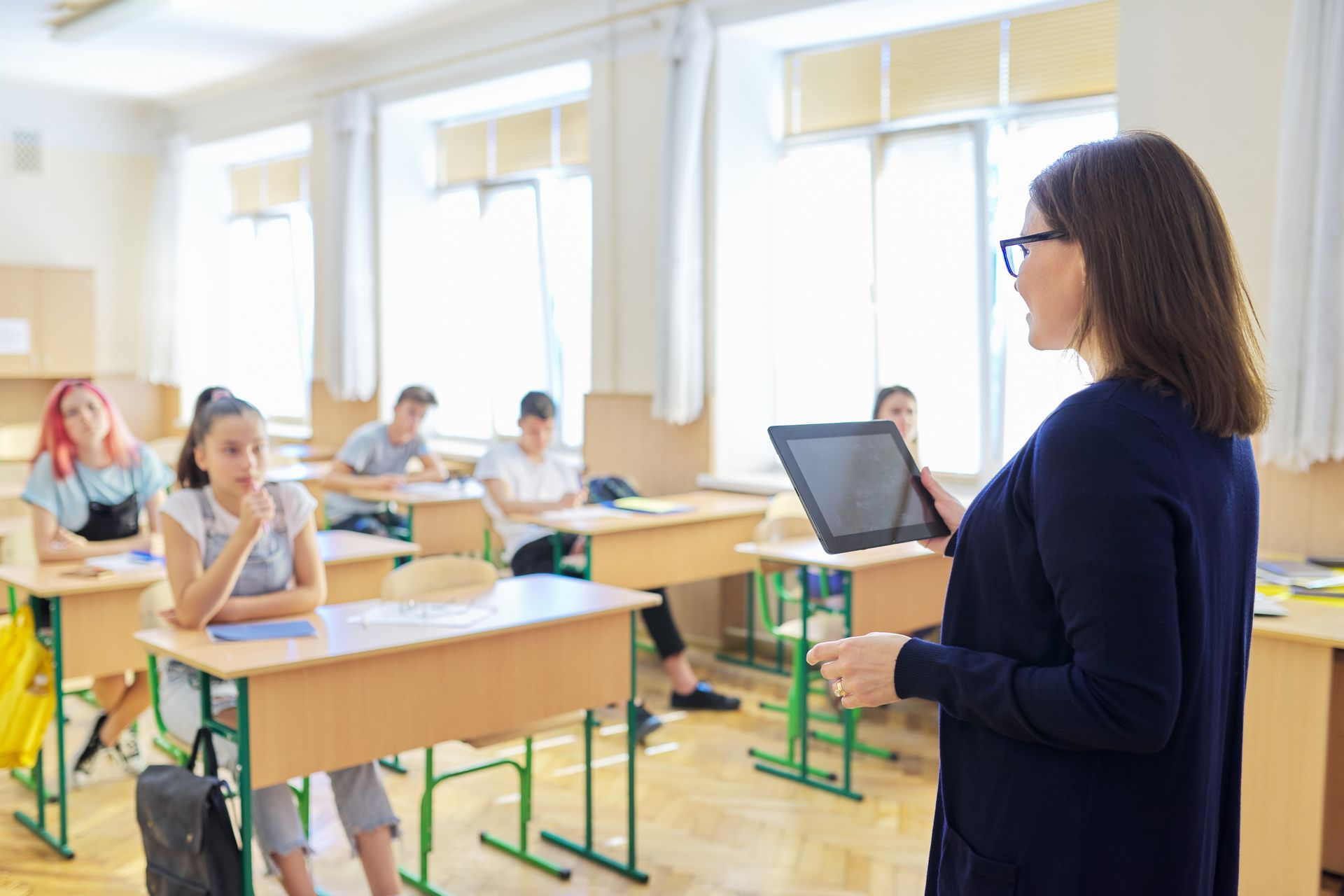 Teacher in glasses holds a tablet, addressing students at desks in a brightly lit classroom.