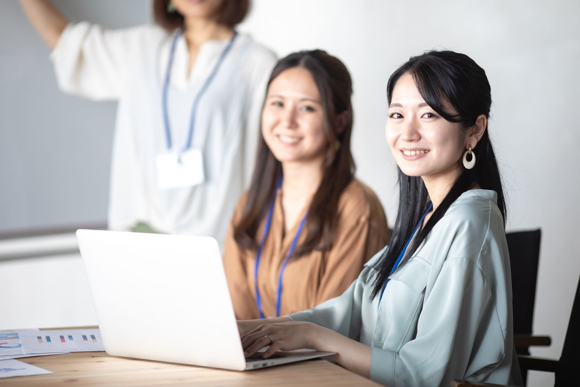 Three women smiling in an office setting; one works on a laptop while others observe a presentation.