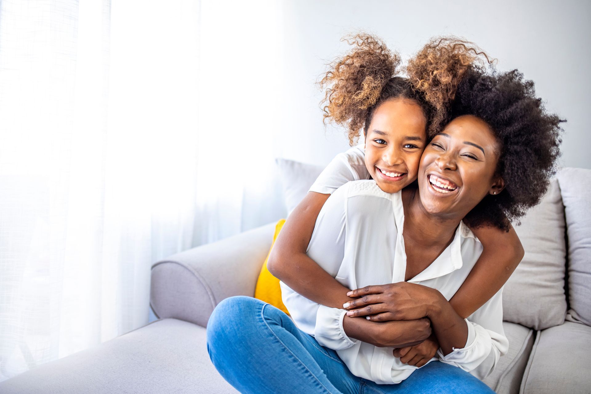 Mother and daughter hugging on a couch; both smiling broadly, light-filled room.