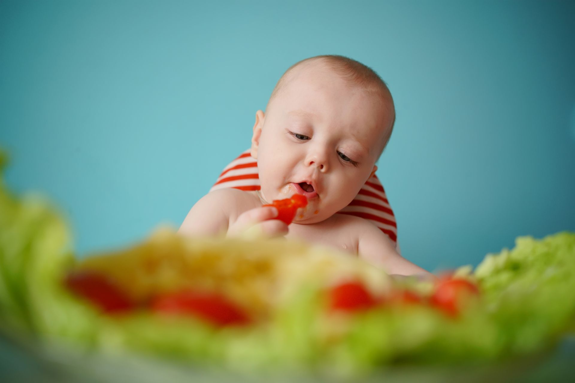 Bebé comiendo un tomate rojo, rodeado de lechuga verde, sobre un fondo azul.