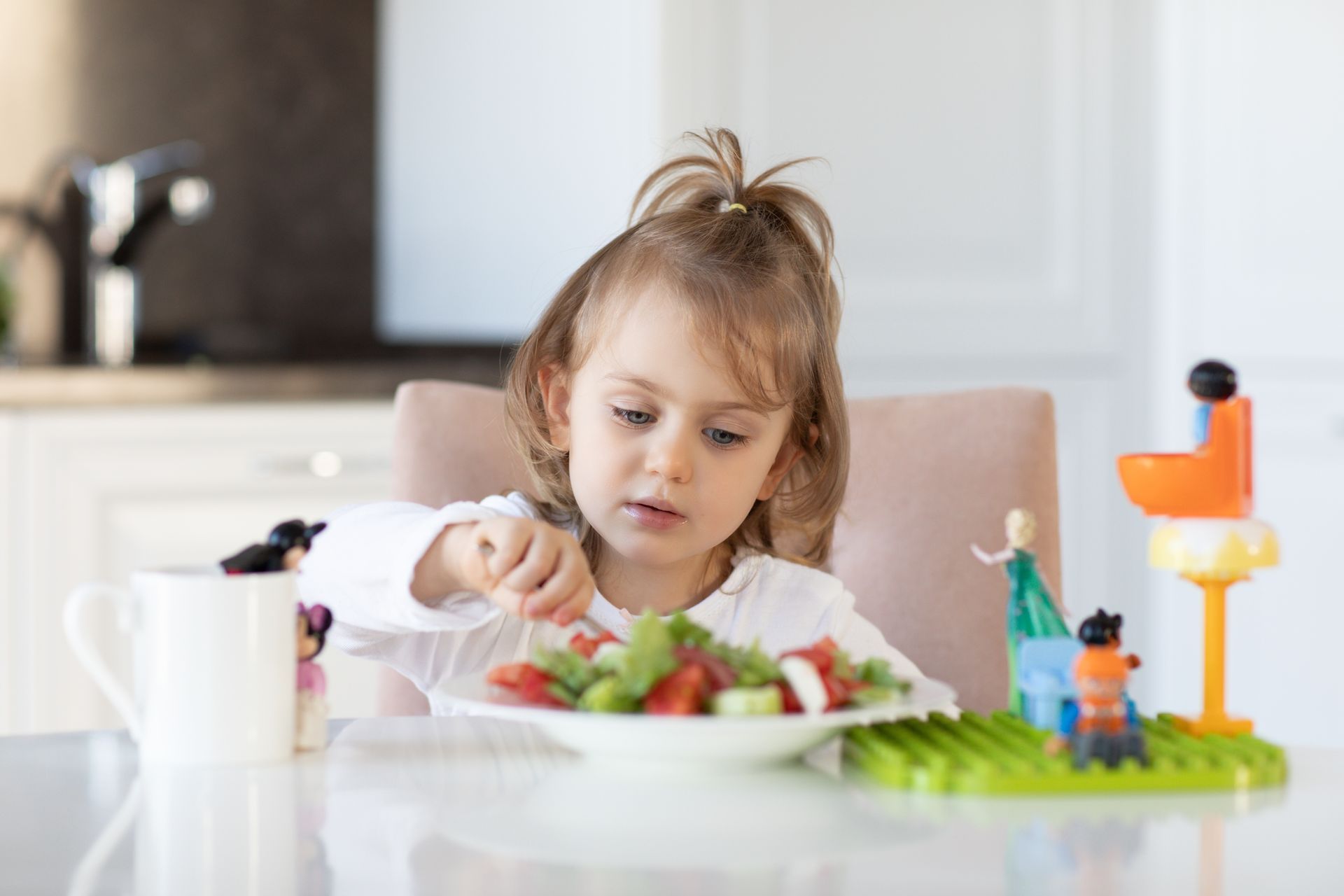 Niña comiendo ensalada en una mesa en una cocina; juguetes en la mesa; taza blanca.