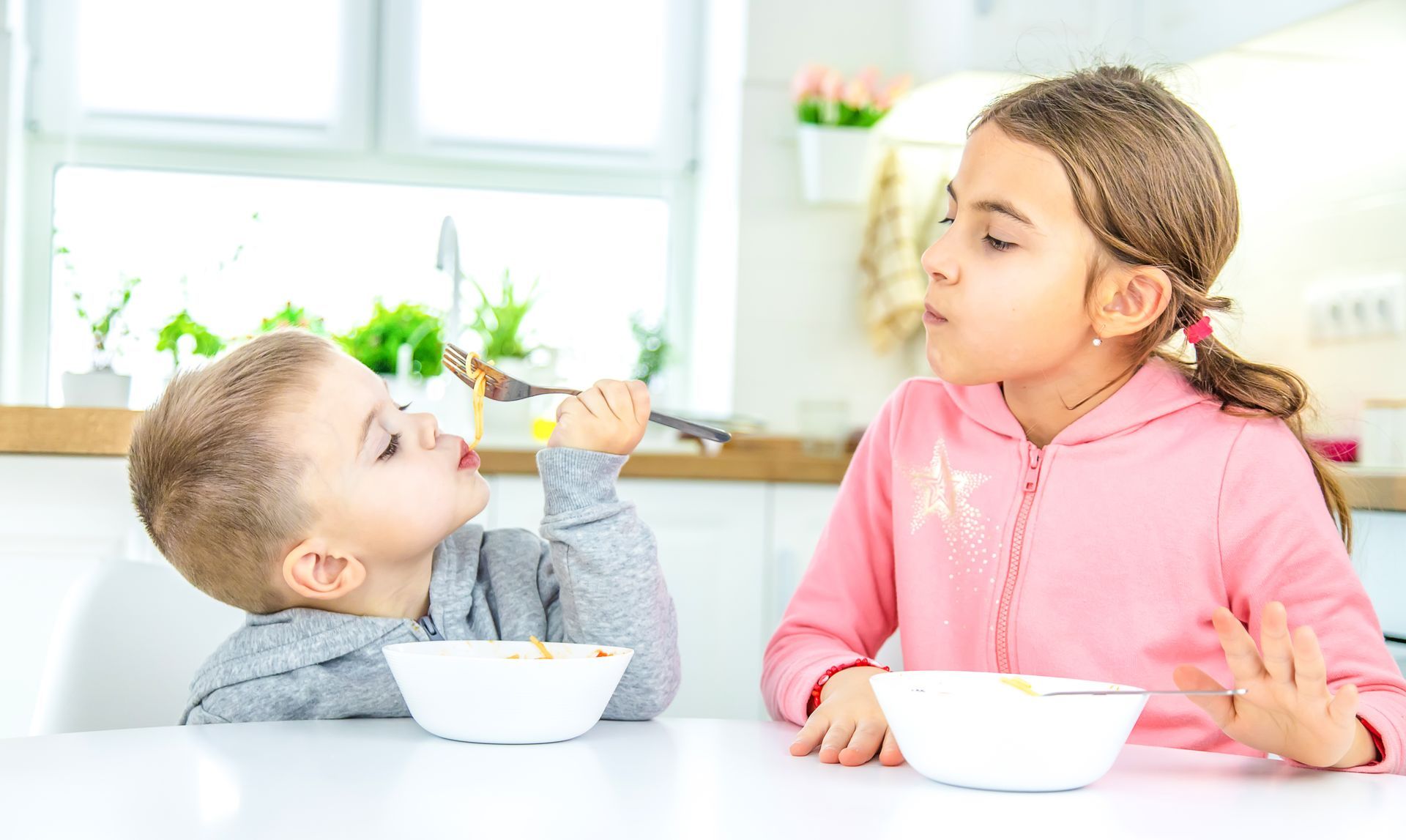 Un niño le da de comer a una niña en una mesa de cocina. La niña parece disgustada.