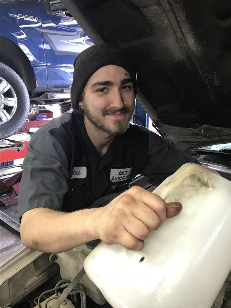 Mechanic in a dark beanie smiles, holding a white reservoir tank under a car's hood in a garage. | Joe's Auto and Tire 