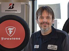 Man in auto shop smiling, standing by Firestone and Bridgestone tires. | Joe's Auto and Tire 