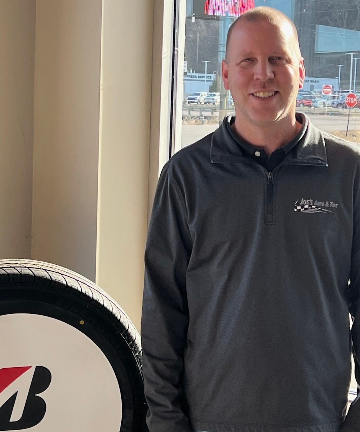 Man smiling in a gray jacket stands near a Bridgestone tire inside a shop. | Joe's Auto and Tire 