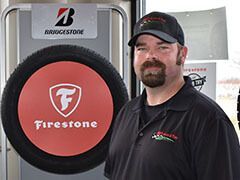 Man in a cap and work shirt stands by a Firestone tire display, beneath a Bridgestone sign. | Joe's Auto and Tire 