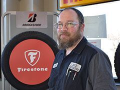 Man in a mechanic uniform, standing near a Firestone tire display and Bridgestone sign. | Joe's Auto and Tire 