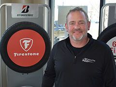 Man smiling in front of a Firestone tire display, wearing a black shirt with a logo. | Joe's Auto and Tire 