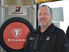 Man in a black jacket smiling, standing in a tire shop in front of Firestone and Bridgestone logos. | Joe's Auto and Tire 