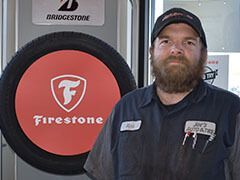 Man in auto shop uniform stands near a Firestone tire sign, looking at the camera. | Joe's Auto and Tire 