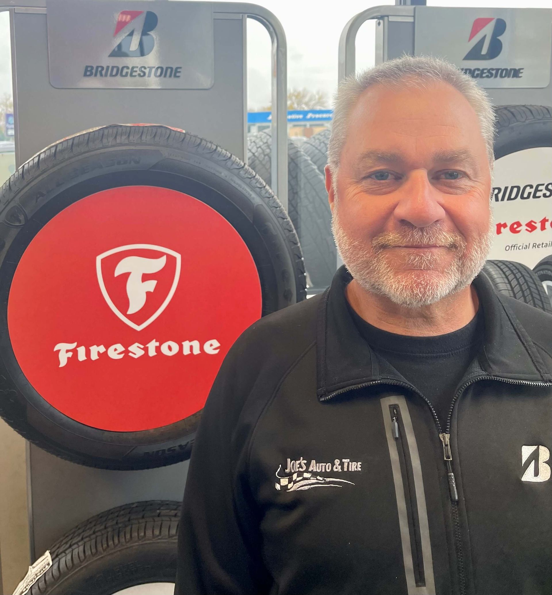 Man in a black jacket smiles in front of Firestone and Bridgestone tire displays. | Joe's Auto and Tire 