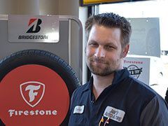Man in blue uniform smiles, stands by a Firestone tire display and Bridgestone sign inside a tire shop. | Joe's Auto and Tire 
