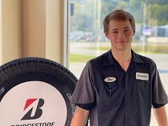 Man in mechanic uniform stands next to a Bridgestone tire, smiling by a window. | Joe's Auto and Tire 