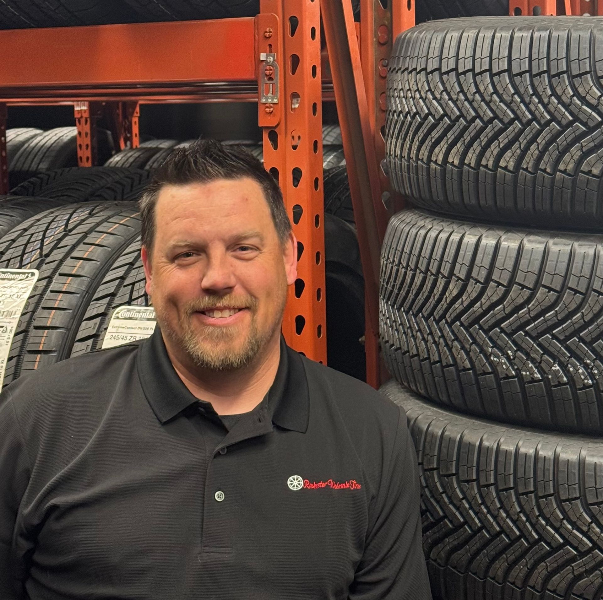 Man in black shirt smiles, standing in a tire store with stacked tires in background. | Joe's Auto and Tire 