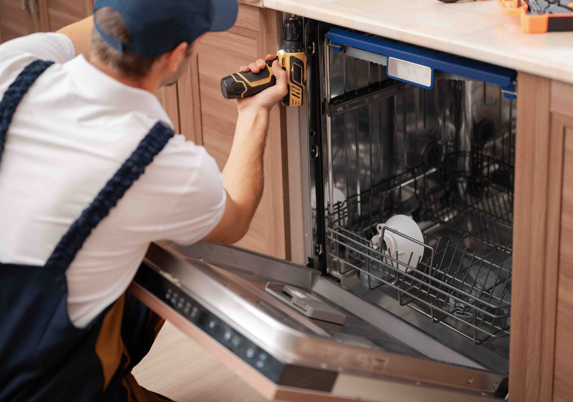 A man is fixing a dishwasher in a kitchen with a drill.
