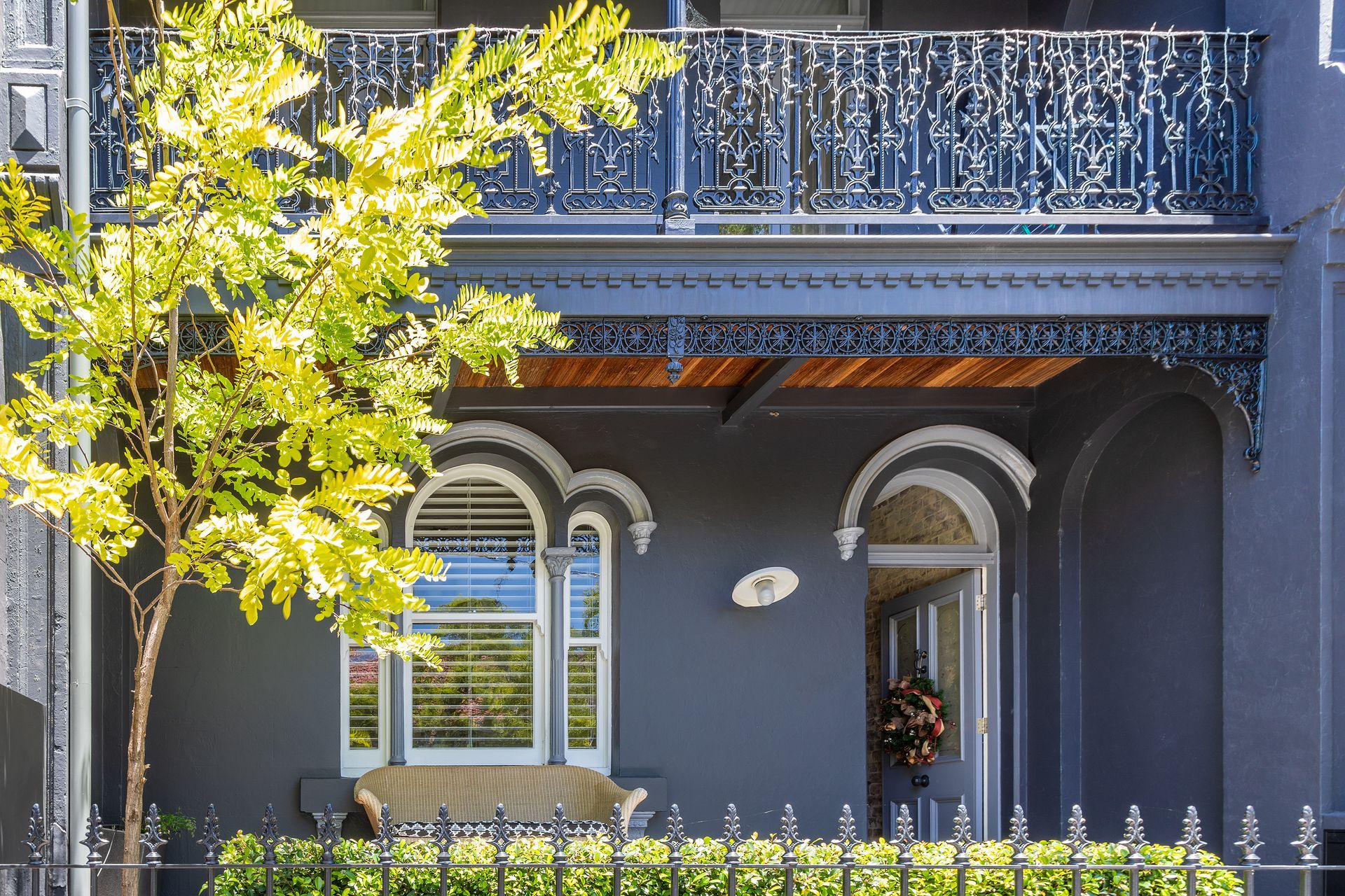 Gray Victorian house facade with ornate balcony, arched windows, and a leafy tree in front