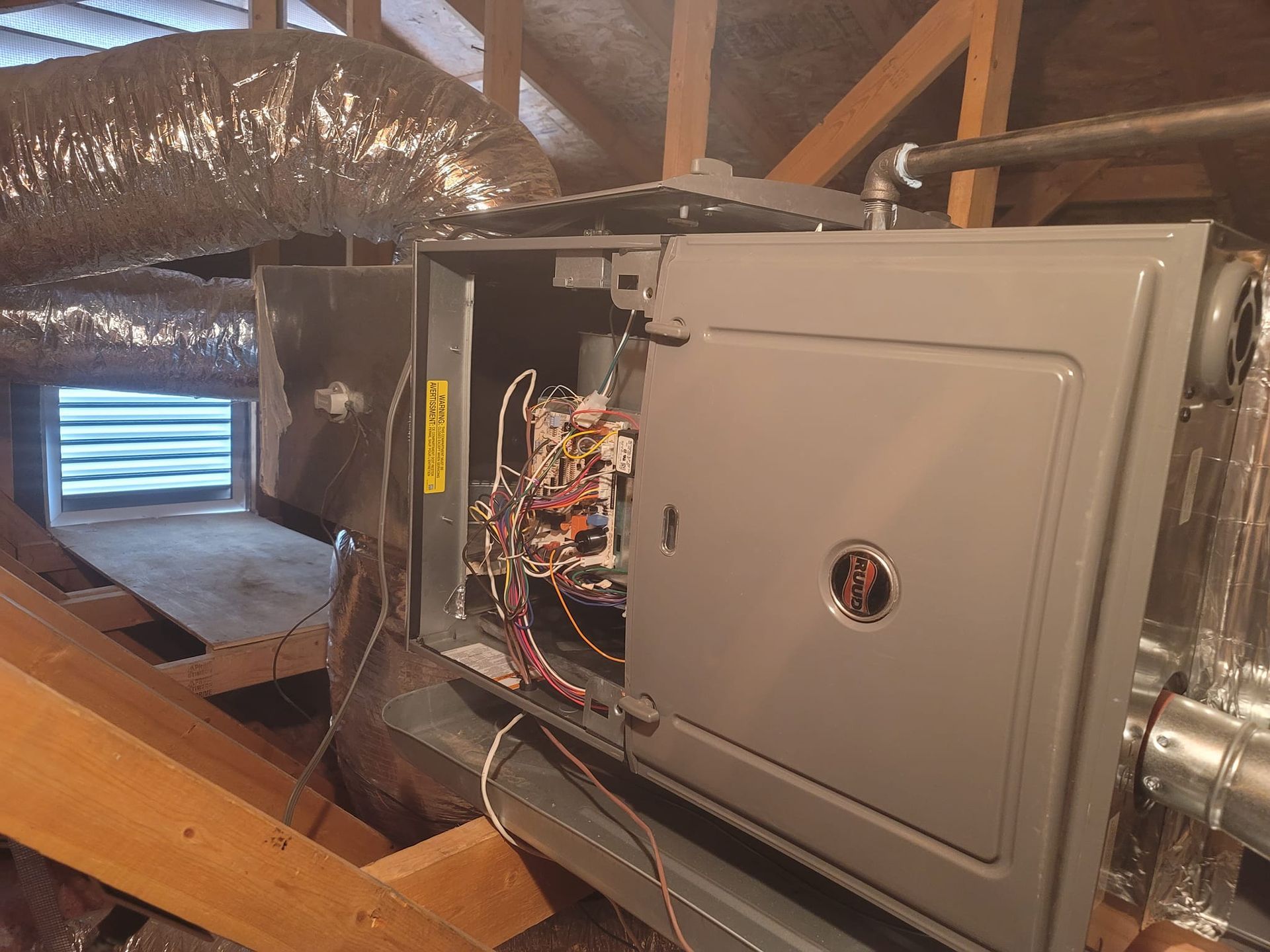 HVAC unit in an attic, partially open, revealing wiring and components. Ductwork and rafters visible.