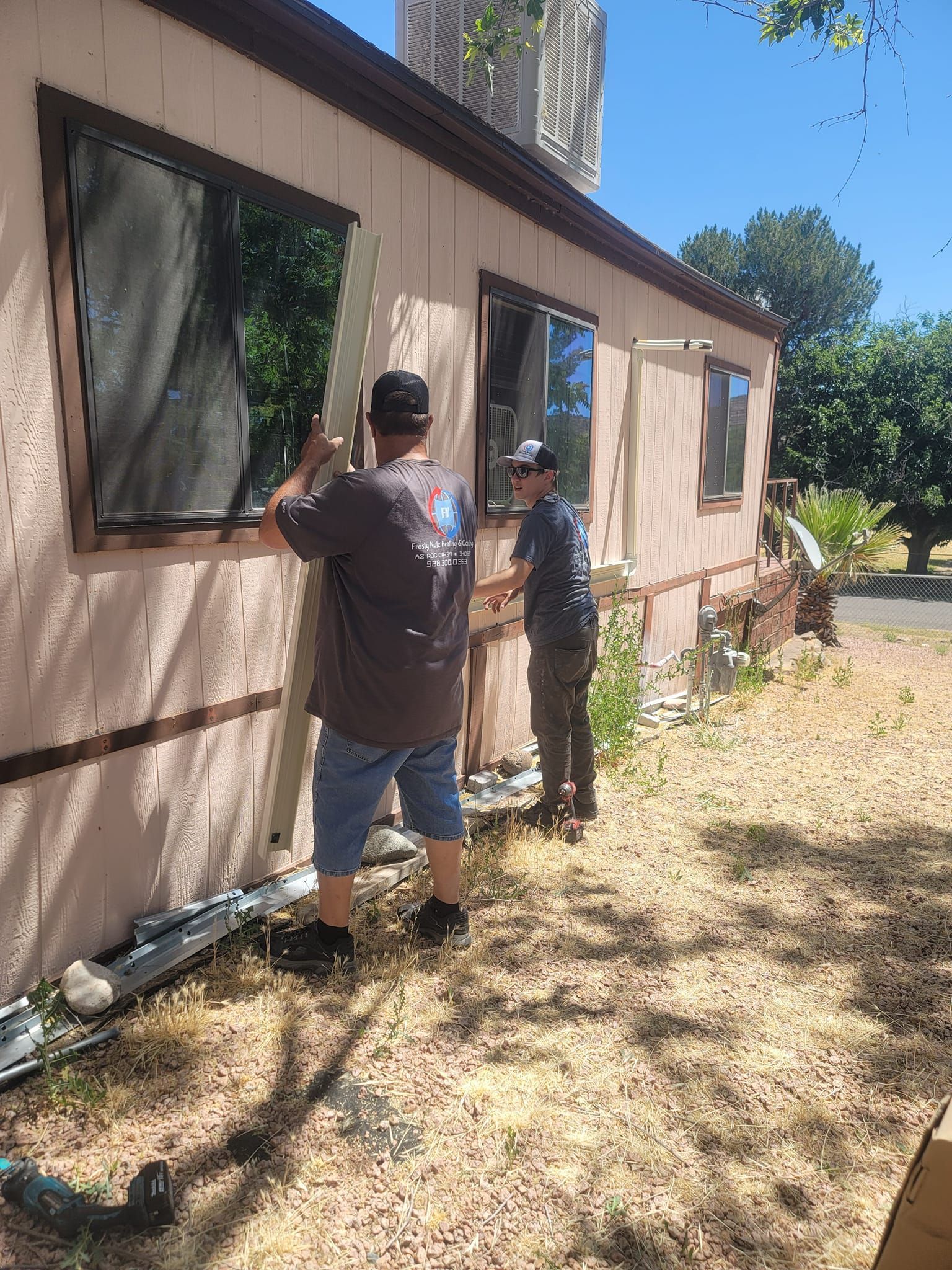 Two people working on the exterior of a tan mobile home; one holds a long board. Sunny day.