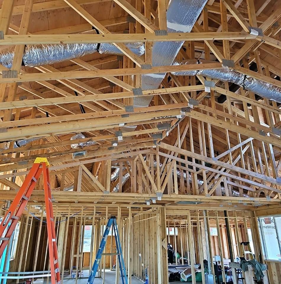 Interior of a house under construction. Wooden framing with rafters and metal ductwork. Two ladders are present.