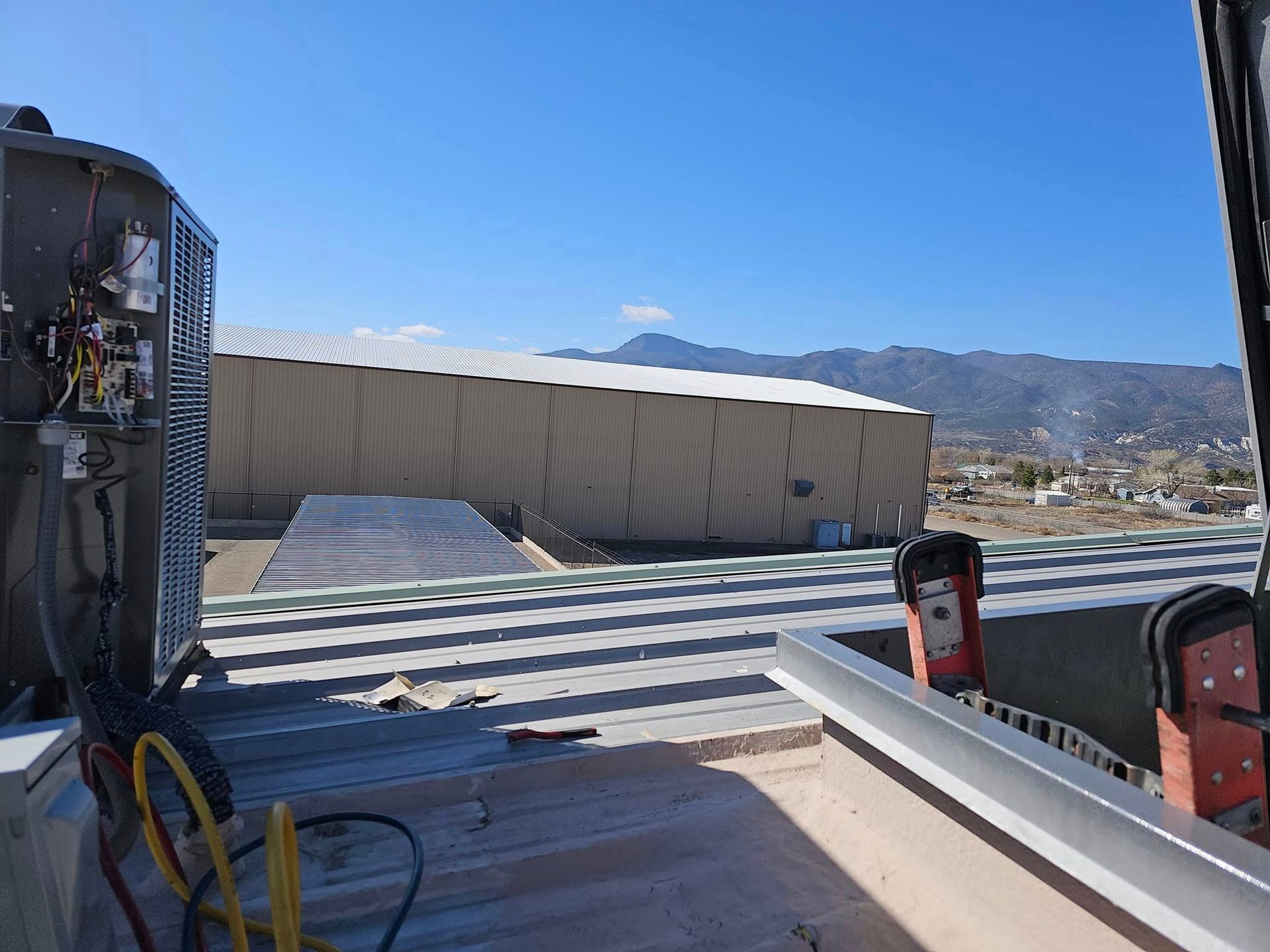 Rooftop view with industrial equipment, a large building, mountains, and a clear blue sky.