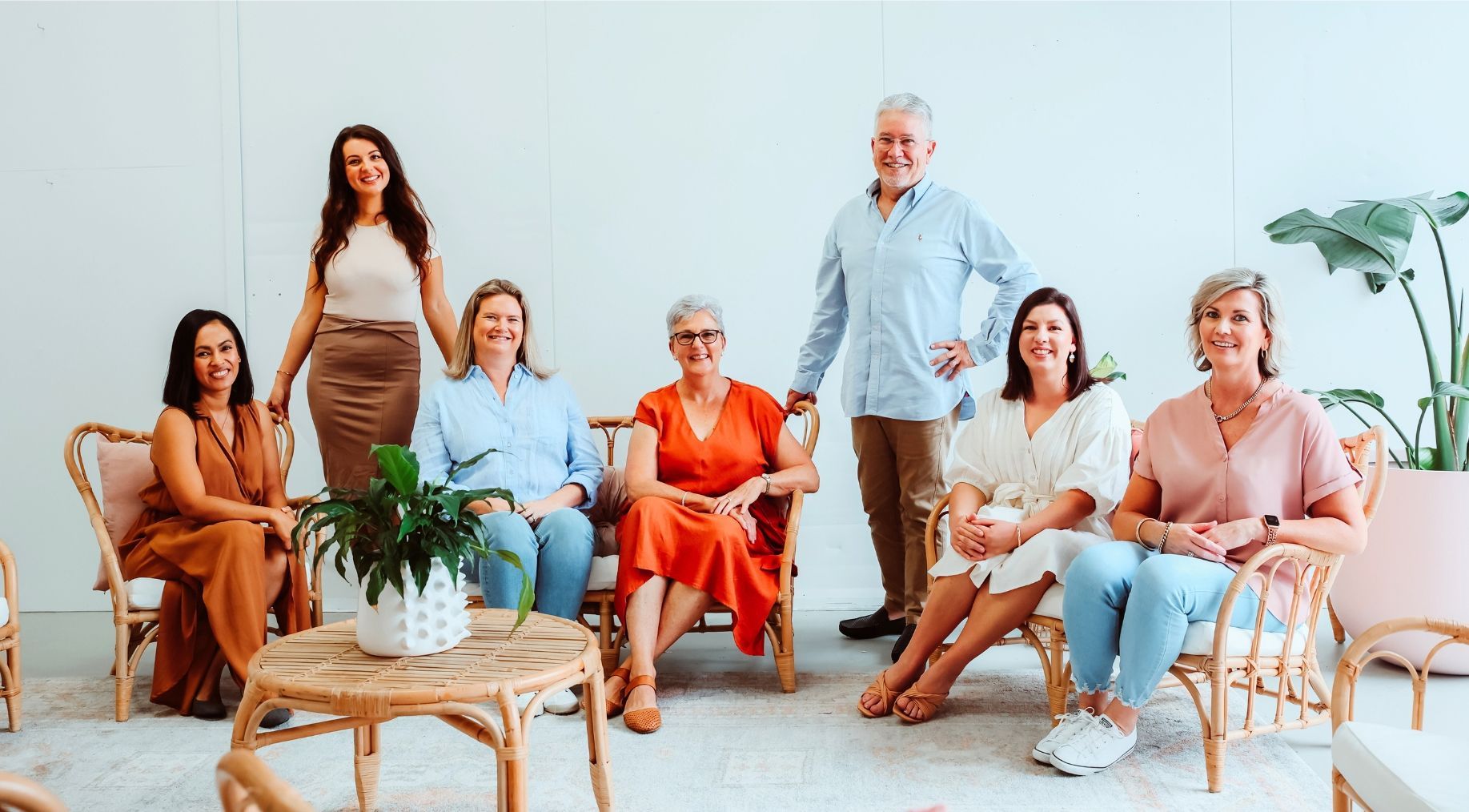 Group of people in chairs and standing, in a well-lit room. Smiling, with plants and neutral background.
