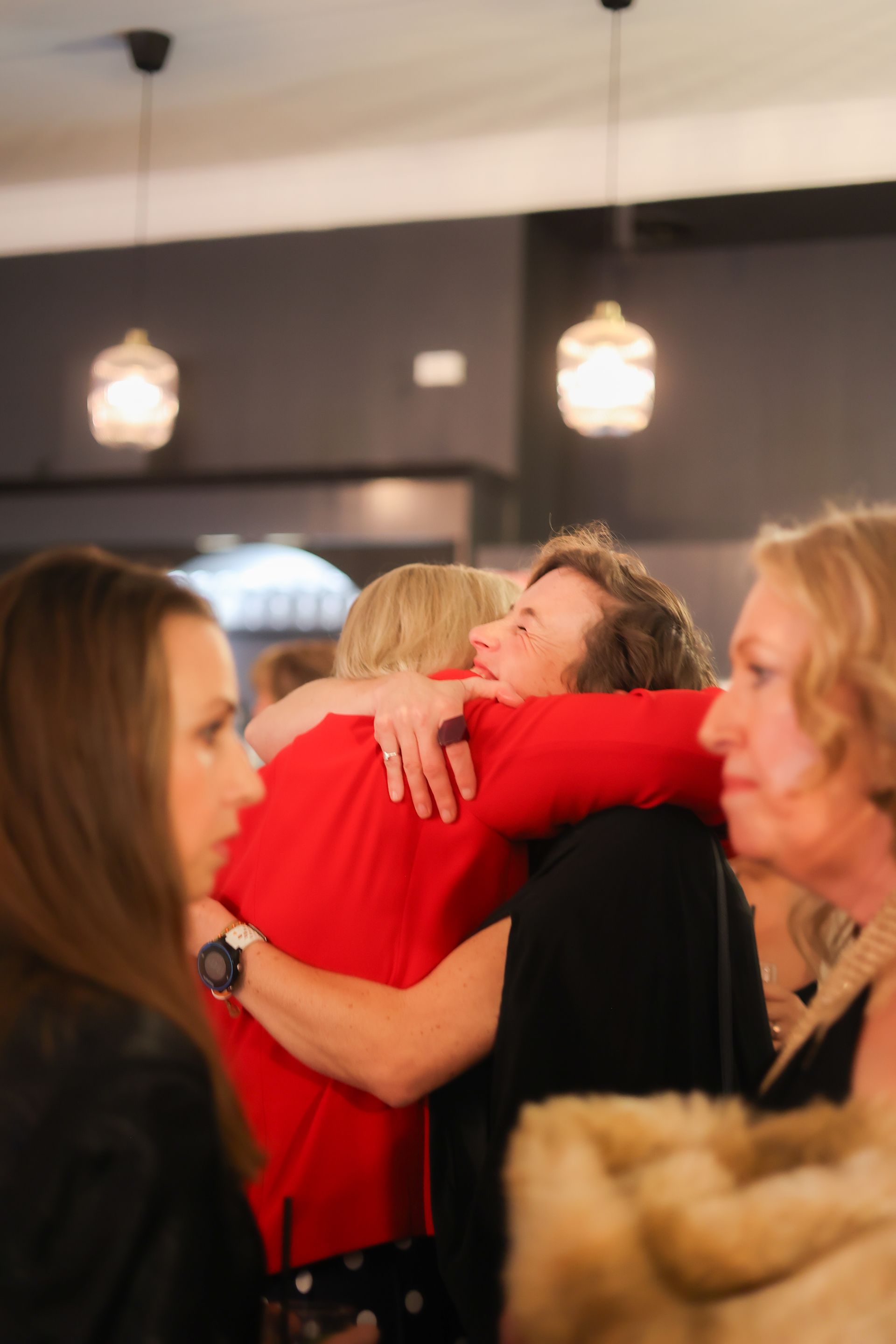Two people embracing indoors, one in red, others watch.