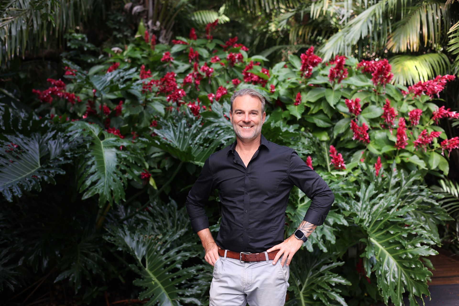 Man in black shirt and gray pants smiles, posing in front of lush tropical foliage with red flowers.