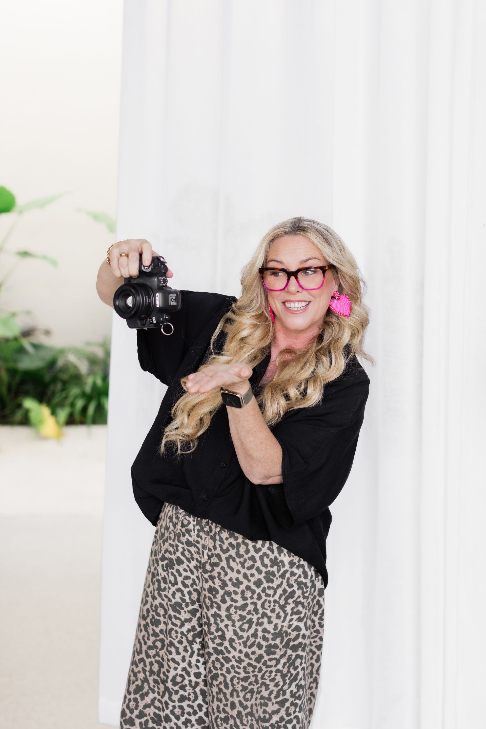 Woman holding a camera, gesturing. Wearing glasses, pink earrings, black top, animal print pants, in a bright setting.