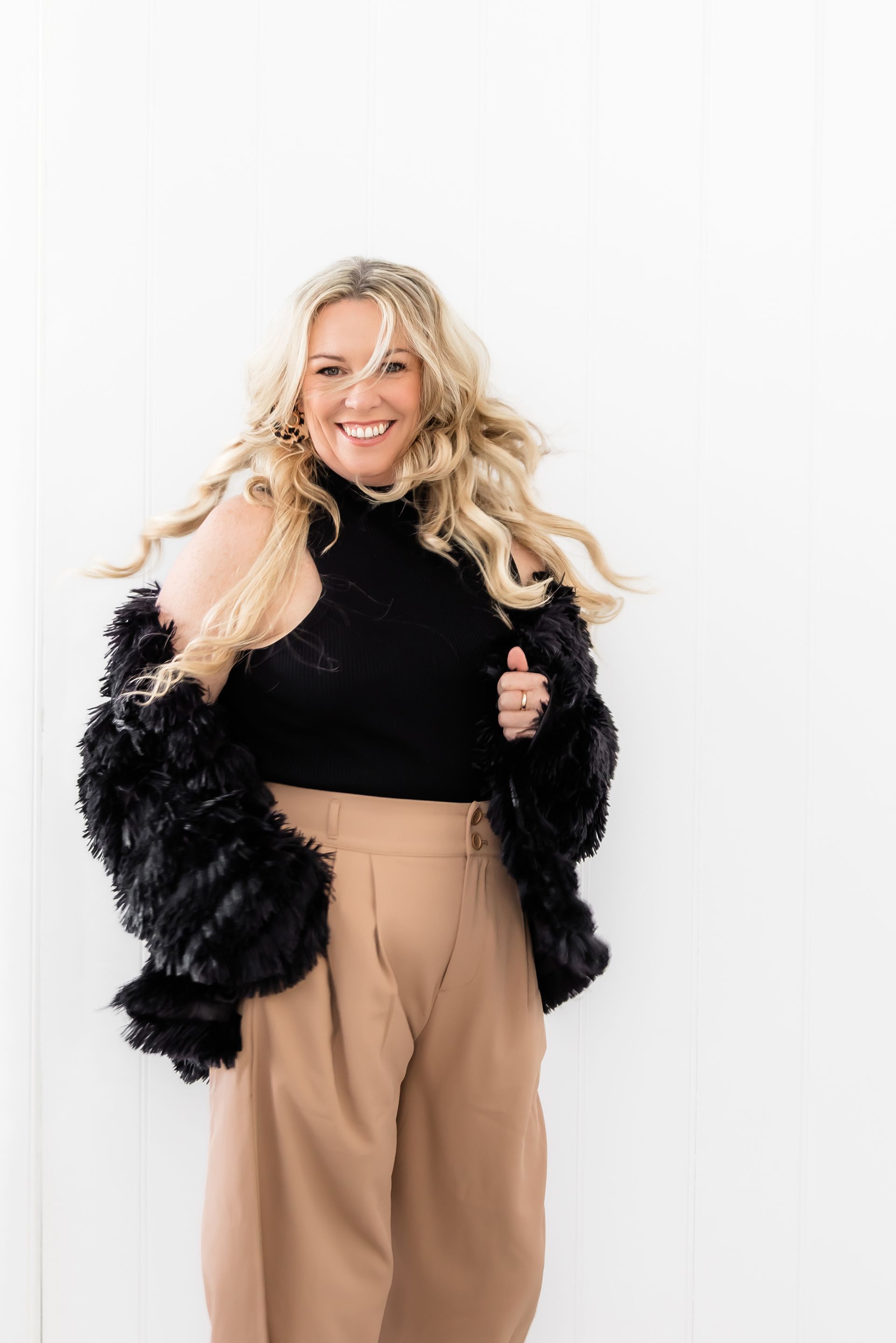 Woman smiles, wearing black top, fuzzy black jacket, tan pants, in front of a white wall.