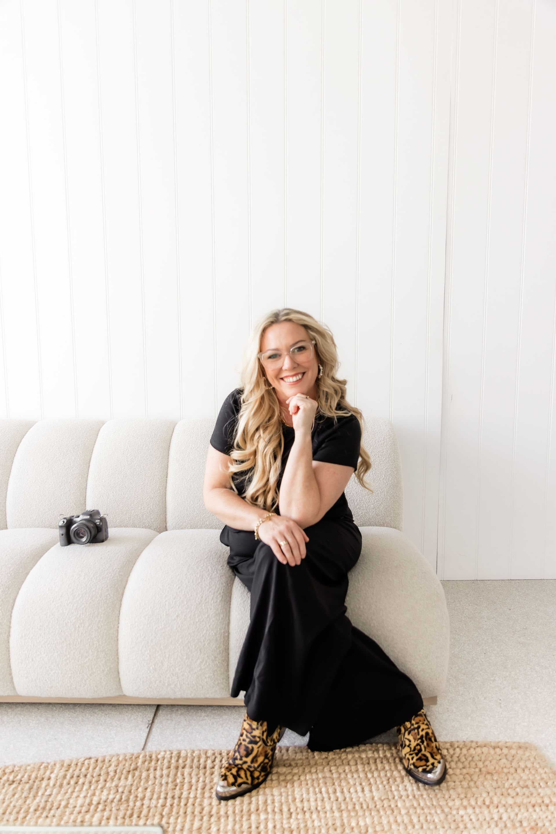 Woman with blonde hair, smiling, seated on a beige couch, wearing black dress and animal print boots.
