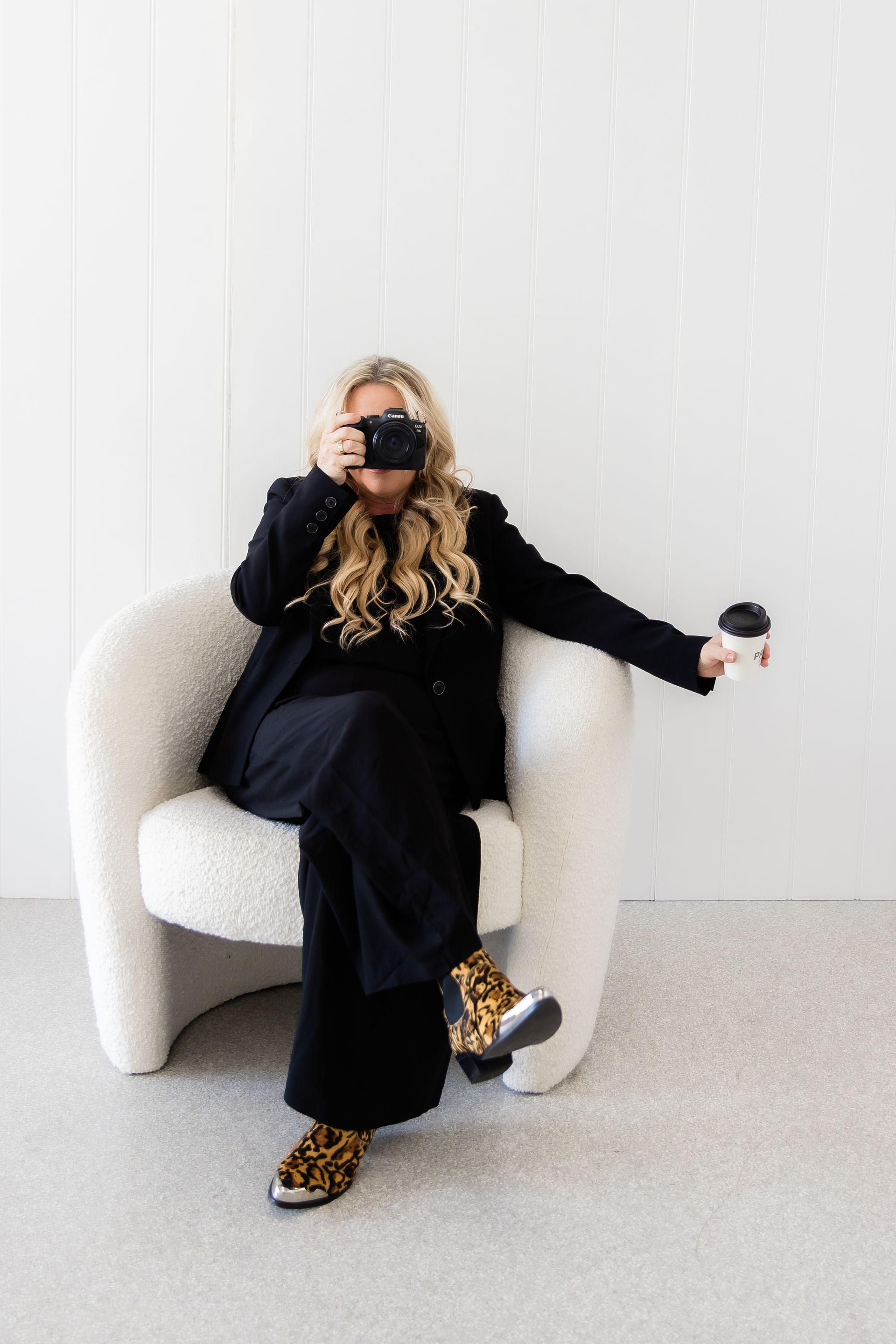 Woman in black suit and leopard print shoes sits in a white chair, taking a photo with a camera.